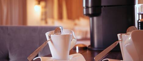Una preparación de café con una taza y platillo sobre una mesa, una cafetera y un sofá al fondo.