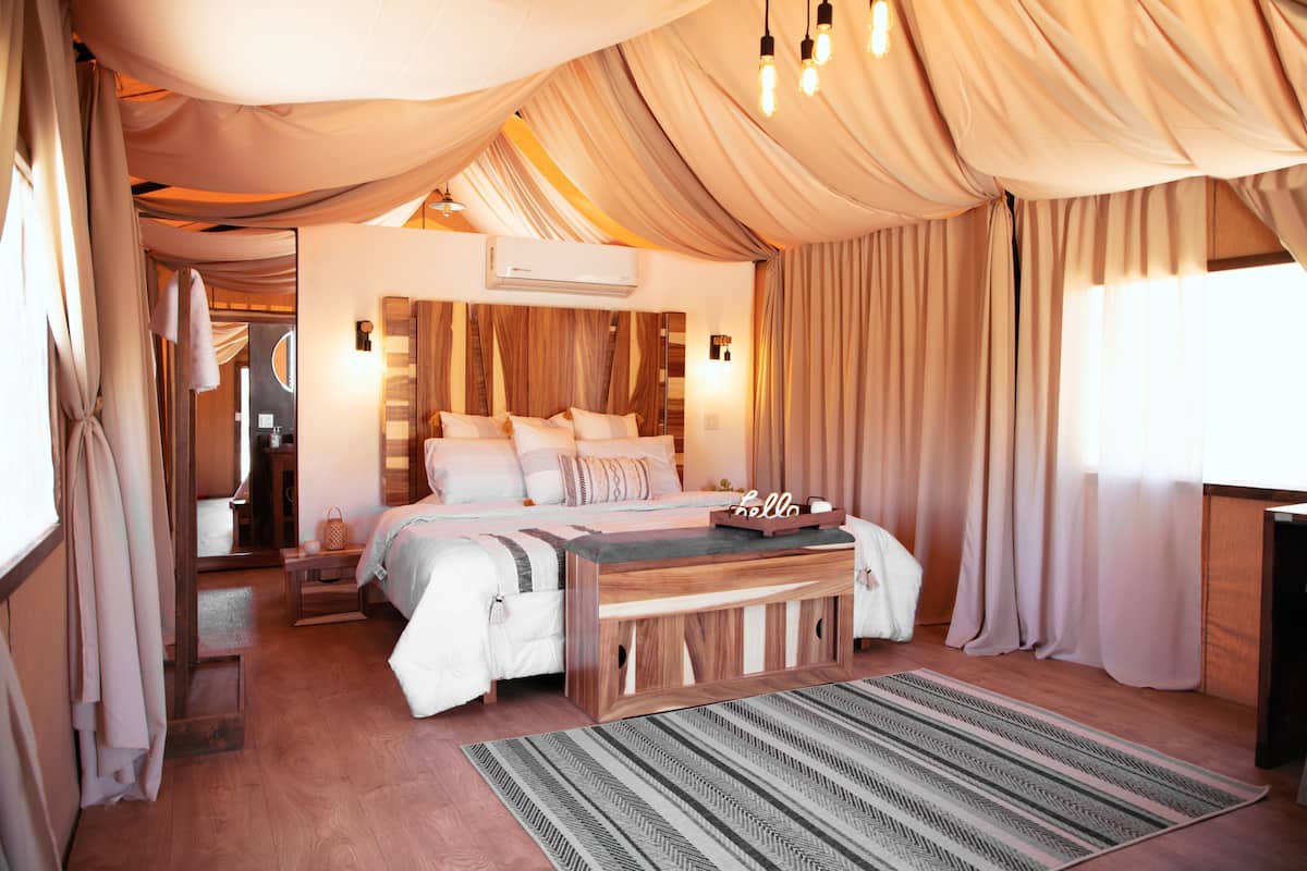 A bedroom with a bed, bedside tables, a bench, a rug, curtains, and an air conditioning unit.