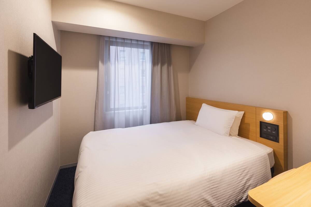Habitación de hotel con cama, televisor de pantalla plana montado en la pared, mesita de noche y ventana con cortinas translúcidas.