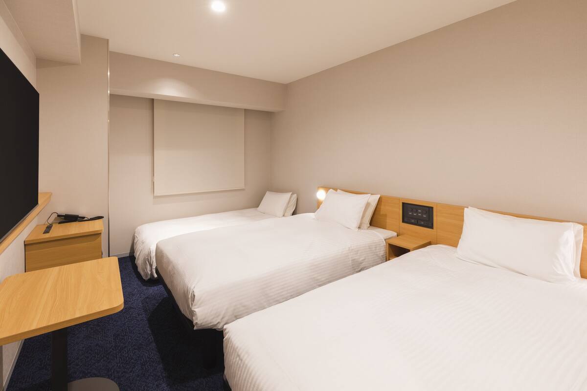 Habitación de hotel con dos camas individuales, un escritorio, una silla, un televisor y un panel de control en la pared.