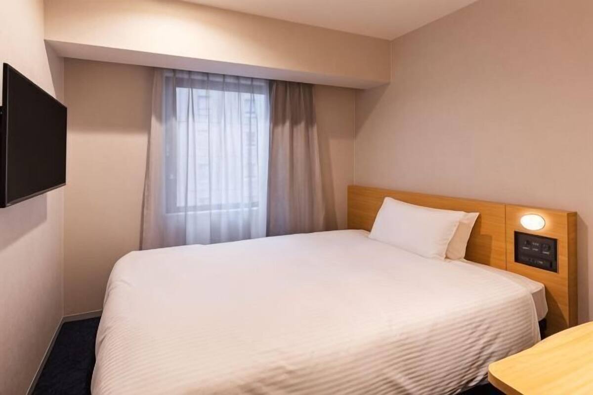 Una habitación de hotel con cama, televisión instalada en la pared, ventana con cortinas y mesita de noche.
