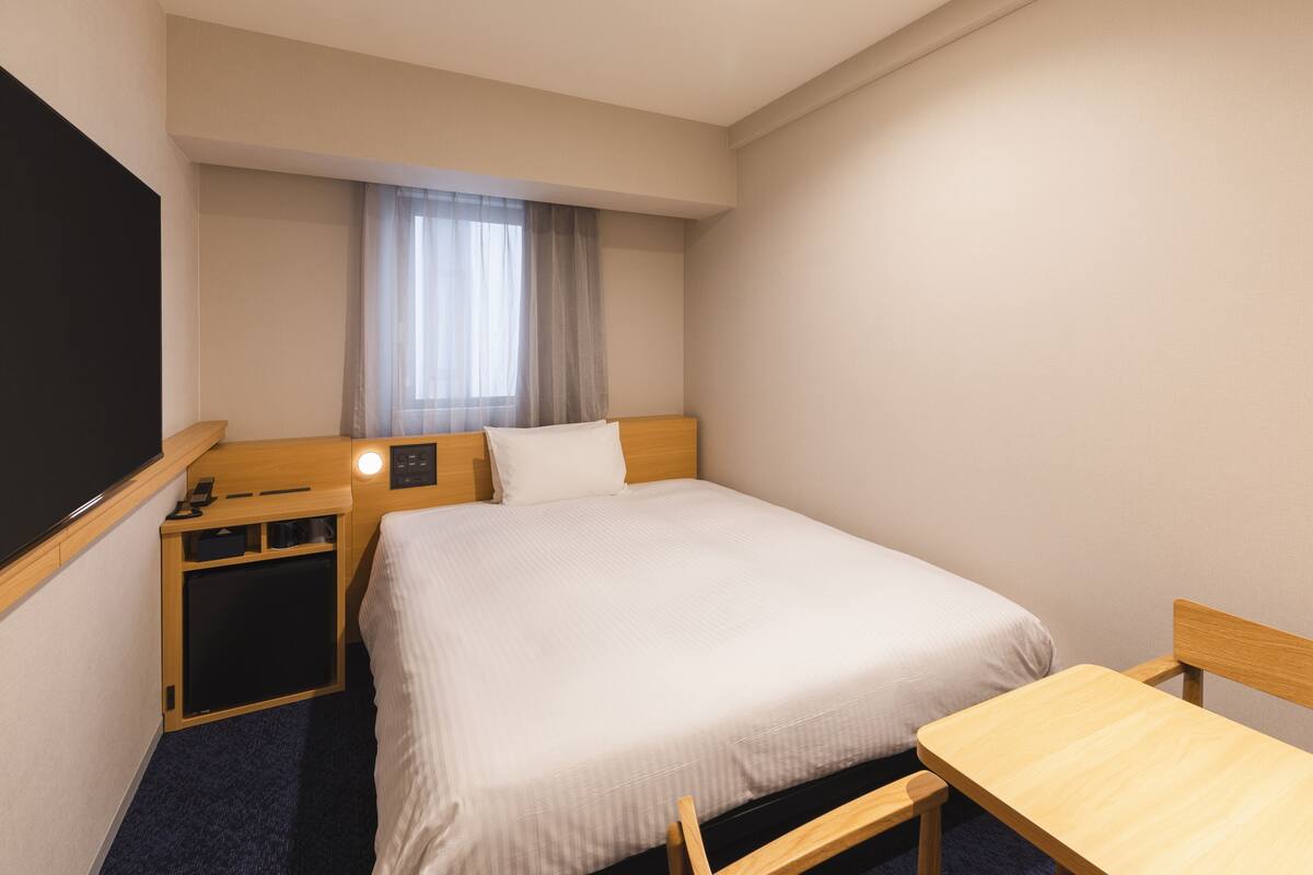 Habitación de hotel con una cama individual, una mesita de noche, un pequeño escritorio con una silla, un televisor y una ventana con cortinas.