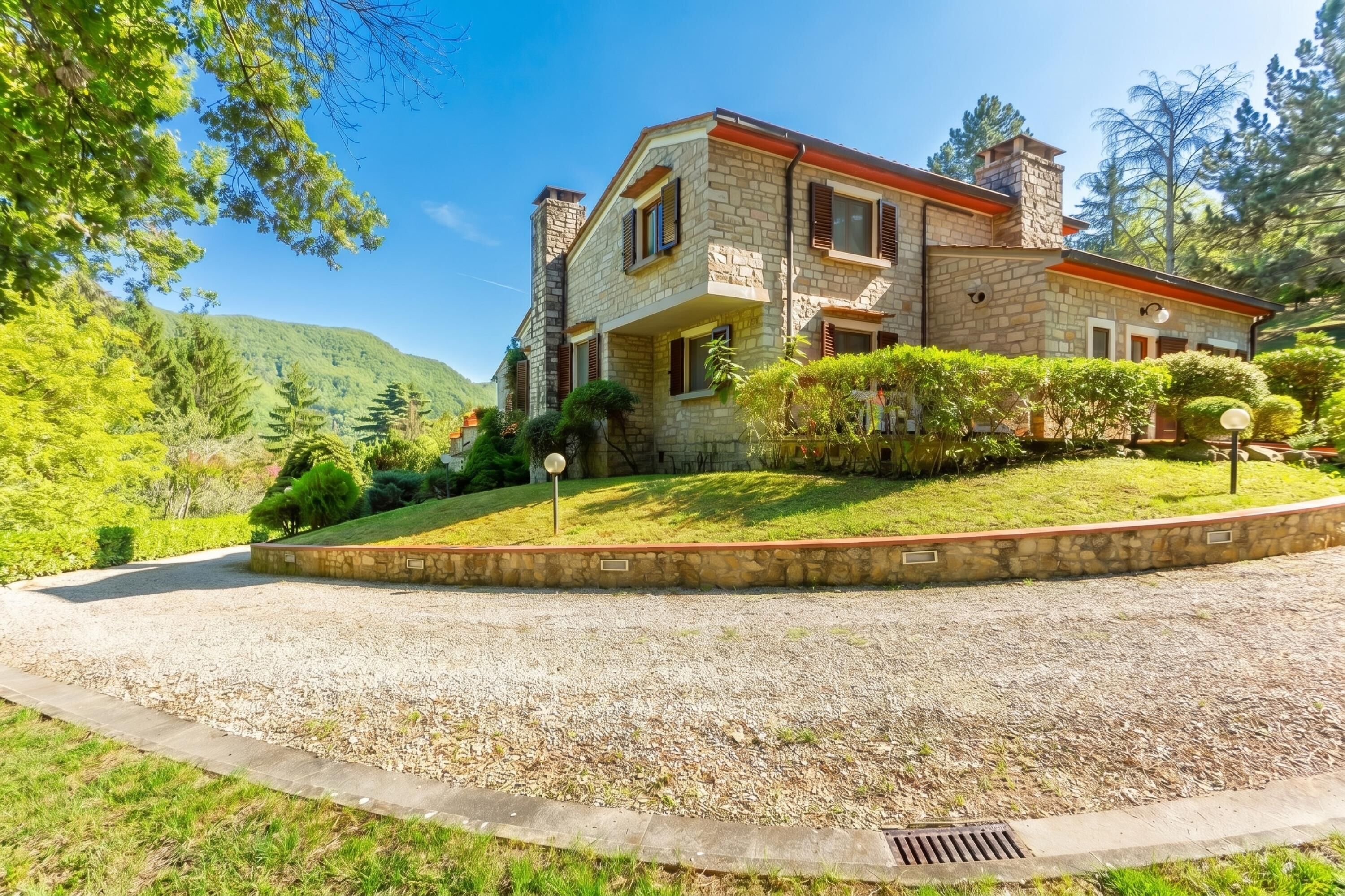Una casa in pietra a due piani con vialetto in pietra, immersa nel verde e con le montagne sullo sfondo.
