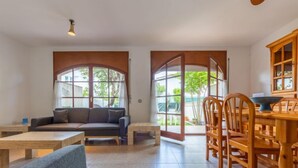 A spacious living area with a sofa, coffee table, and dining table with chairs. Large windows provide natural light and views of the outdoors.