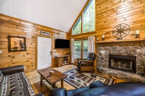 Cozy cabin-style room with a stone fireplace, television, and large window offering a view of the outdoors.