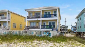 A multi-story residential building with balconies, a wooden fence, and a sandy beach in the foreground. - Oceanfront Escape: 5BR Beach House w/ Pool for 12 (Gulf Shores)
