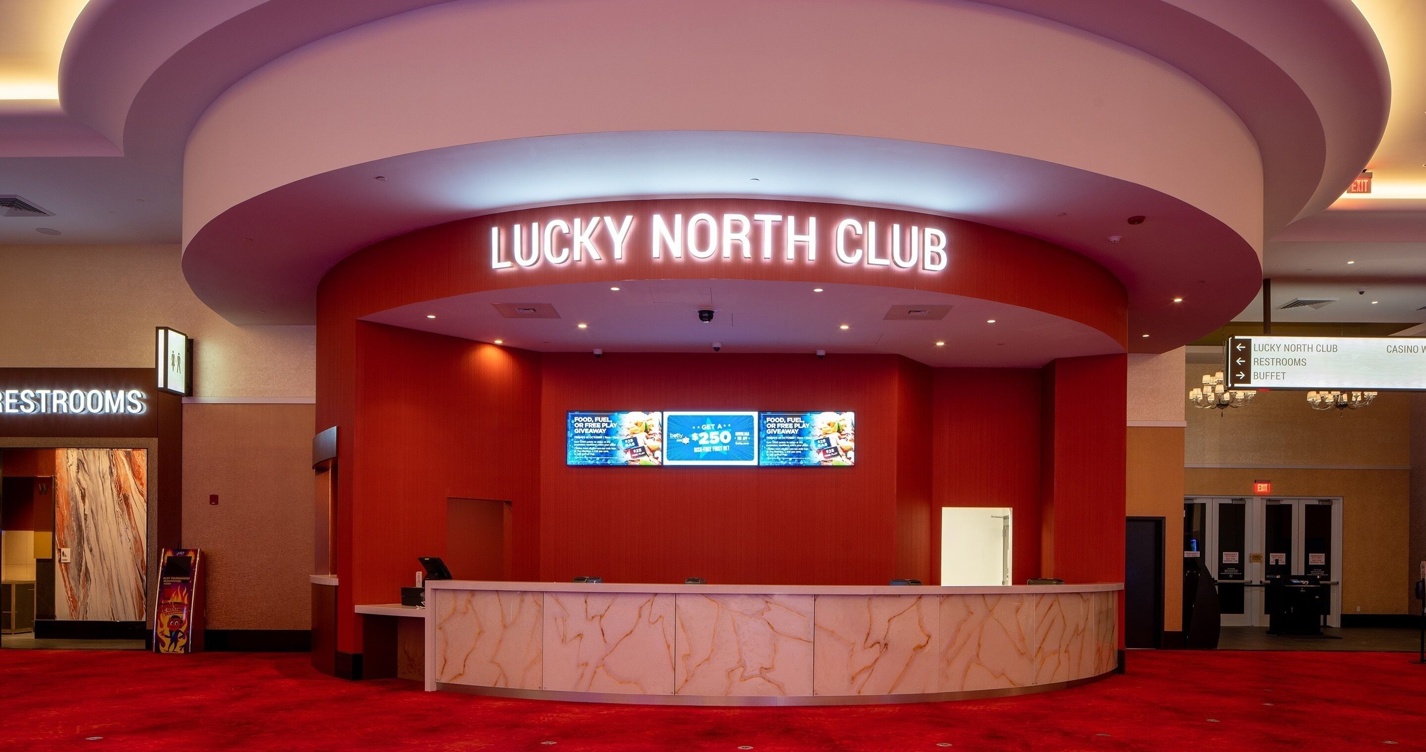 A reception desk at the Lucky North Club with a marble design, a digital screen above displaying information, and a red carpeted floor.