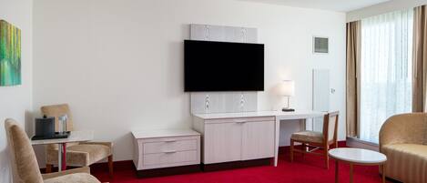 A hotel room with a flat-screen TV, a desk, a chair, a small table, and a red carpet.