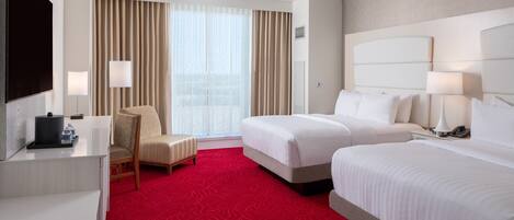 A hotel room with two beds, a desk with a flat-screen TV, a chair, and a large window with curtains.