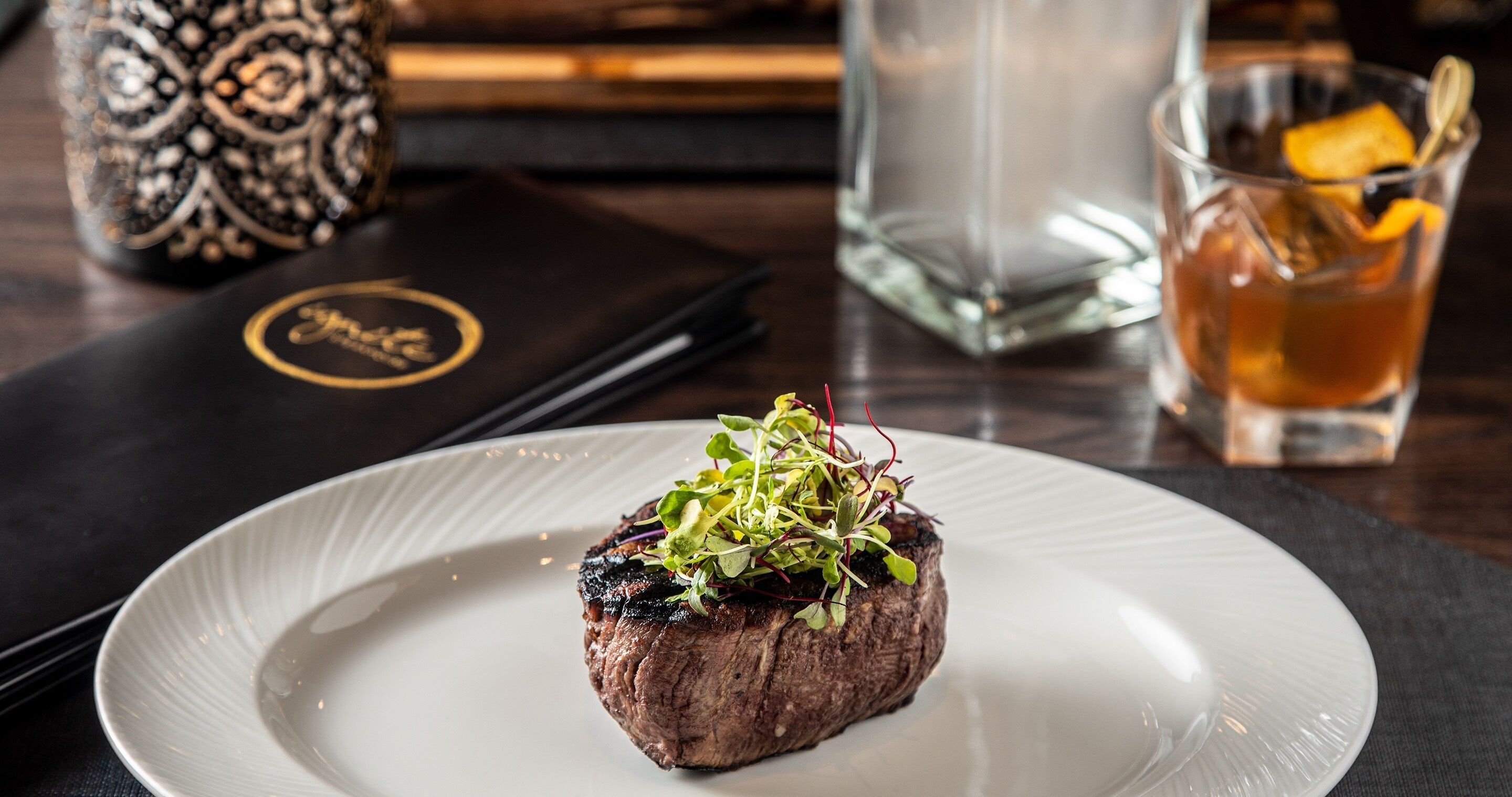 A plate with a steak topped with greens, a glass of tea, and a menu.