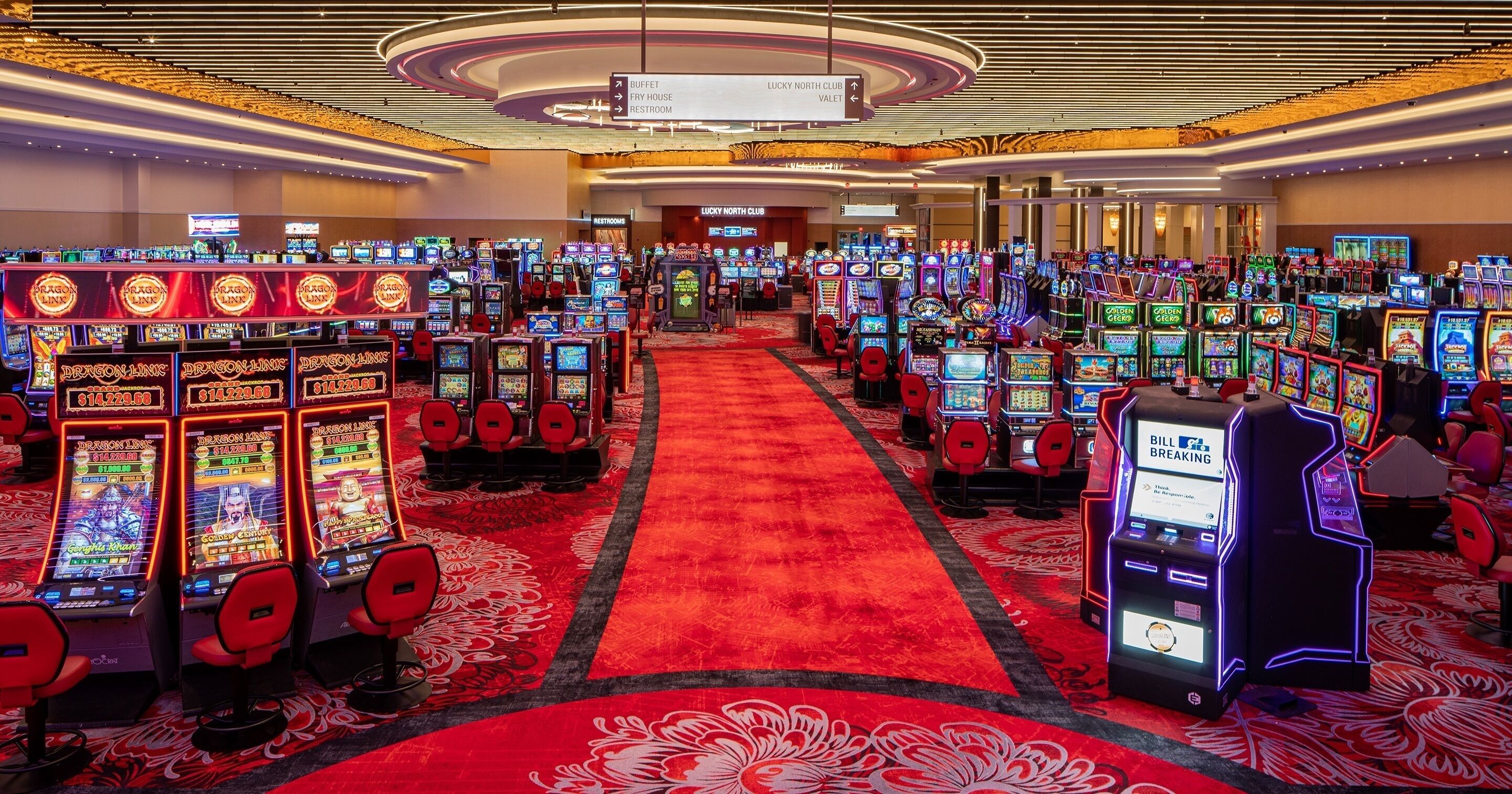 A casino with multiple slot machines arranged in rows, a red carpet, and directional signs for the buffet, lucky north club, and restroom.