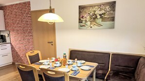 En matplats med ett träbord dukat för fyra personer, omgiven av bekväma stolar. En tavla med blommor hänger på väggen, och en lampa syns ovanför bordet.