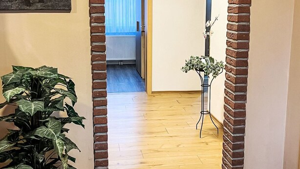 En hall med en tegelbåge som leder in till ett rum med ett element och en dörr. På vänster sida står en krukväxt och på höger sida en vas med blommor.