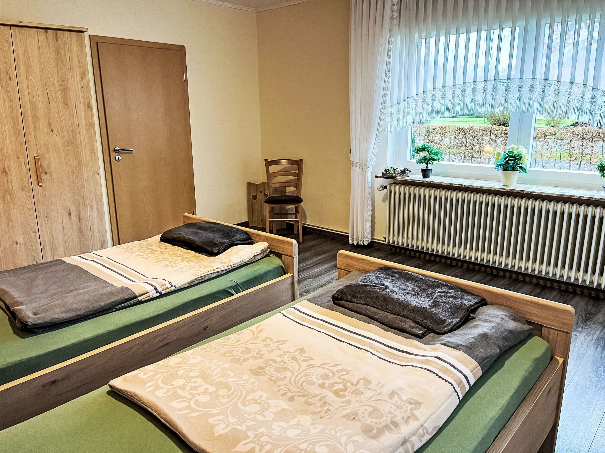A room with two single beds, a wooden wardrobe, a chair, a radiator, and a window with a view of greenery.