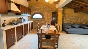 Une cuisine rustique aux murs en pierre, avec des poutres en bois au plafond, une table à manger dressée pour quatre personnes et une cheminée en pierre.
