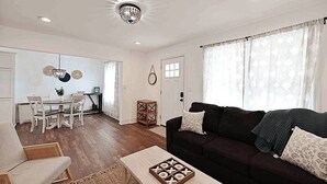 Una sala de estar con un sofá oscuro, una mesa de centro con un juego de mesa, un comedor con una mesa y sillas, y una araña.