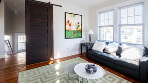 A living room with a dark brown sliding door, a green patterned rug, a black leather sofa with white pillows, a white coffee table, and a painting on the wall. - Contemporary 3-Bedroom Home with Balcony in RVA (Richmond)