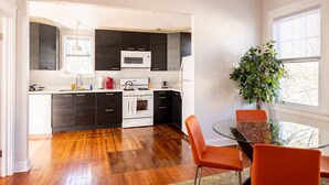 A modern kitchen with dark cabinets, white appliances, and a glass dining table with orange chairs. - Contemporary 3-Bedroom Home with Balcony in RVA (Richmond)