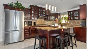 A modern kitchen with dark wooden cabinets, a stainless steel refrigerator, and a granite countertop island with bar stools. - Spacious Luxury 4 bdr, Pool, Hot Tub, Sauna, Patio (Los Angeles)