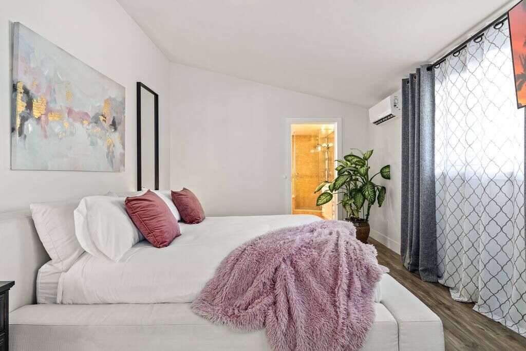 Un dormitorio moderno con una cama blanca, un plaid rosa suave, un cuadro abstracto grande y una planta en maceta.