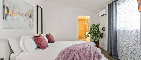 Un dormitorio moderno con una cama blanca, un plaid rosa suave, un cuadro abstracto grande y una planta en maceta.