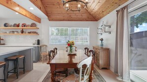A dining area with a wooden ceiling, a central table, and chairs. There are shelves above the counter with dishes and a vase with flowers on the table. - Stylish San Fernando Stay with Designer Kitchen Upgrade (San Fernando)