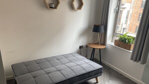 A modern bedroom with a grey sofa bed, a small wooden side table with a lamp, and a window with curtains. - Modern Studio Apt in the heart of Leeds Centre (Leeds)