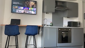 A modern kitchen with a built-in microwave, a dishwasher, and a TV mounted on the wall displaying a streaming service. There are two blue bar stools and a laptop on a wooden table. - Modern Studio Apt in the heart of Leeds Centre (Leeds)