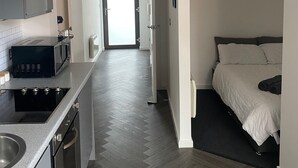 A modern kitchen with a microwave, stove, and sink, leading to a bedroom with a bed and a door to a bright hallway. - Modern Studio Apt in the heart of Leeds Centre (Leeds)