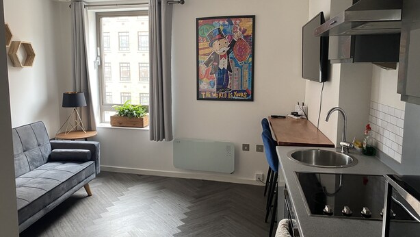 A modern apartment with a grey sofa, a small dining table, a kitchen with a sink and stove, a radiator, and a window with curtains. - Modern Studio Apt in the heart of Leeds Centre (Leeds)