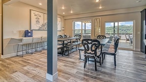 A spacious dining area with a long table and multiple chairs, large windows with a view of greenery, and a ceiling with recessed lighting.