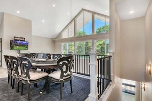A dining area with a high ceiling, large windows, and a TV mounted on the wall.