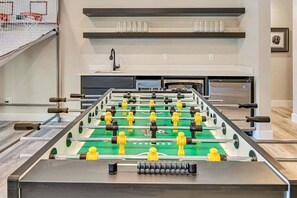 A foosball table in a room with a TV, shelves, and a basketball hoop in the background.