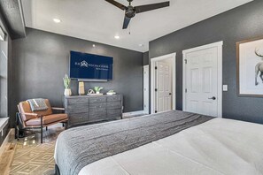A bedroom with a large bed, a dresser, a chair, a ceiling fan, and a TV mounted on the wall.