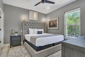A bedroom with a tufted headboard, a bed with white bedding, a bedside table with a lamp, and a window with a view of trees.