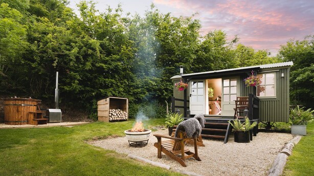 A rustic cabin with a fire pit, wooden deck, and a small shed. - Red Deer Escape - Luxury Cottages (Minehead)