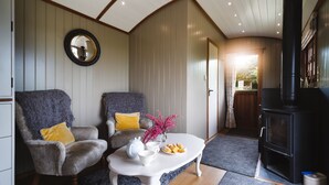 A cozy living room with two armchairs, a white coffee table with a vase and snacks, and a black wood-burning stove. - Hooting Owl Retreat - Luxury Cottages (Minehead)