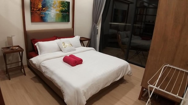 Un lit bien fait, avec du linge de lit blanc et des touches de rouge, une petite table de chevet avec une lampe, et un tableau au-dessus du lit.