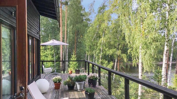 Eine Holzterrasse mit weißem Liegestuhl, Blumentöpfen und Blick auf einen von Bäumen gesäumten See.