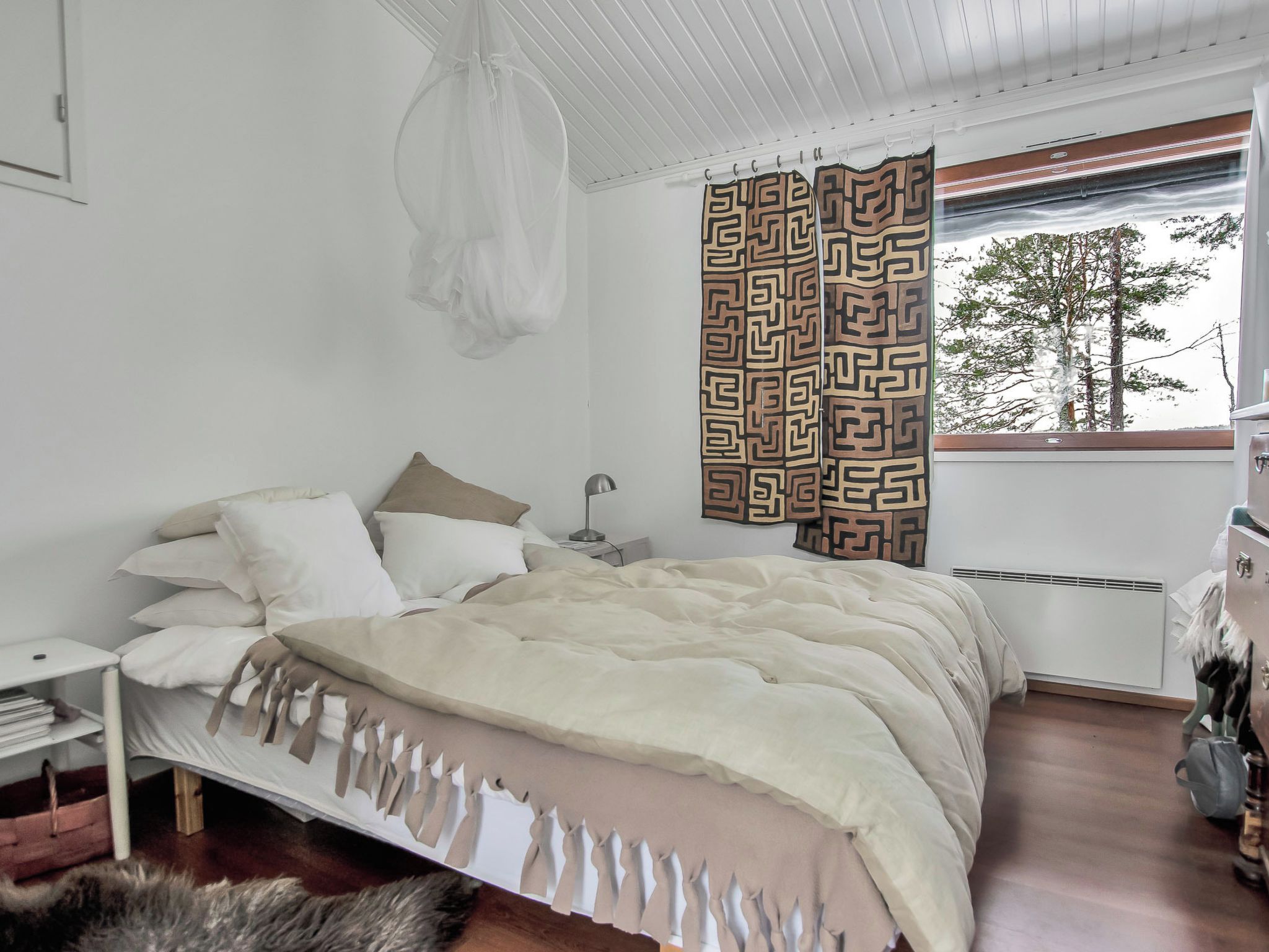 Une chambre à coucher avec un lit, des tables de chevet, une lampe, un rideau décoratif et une vue sur les arbres à l’extérieur.
