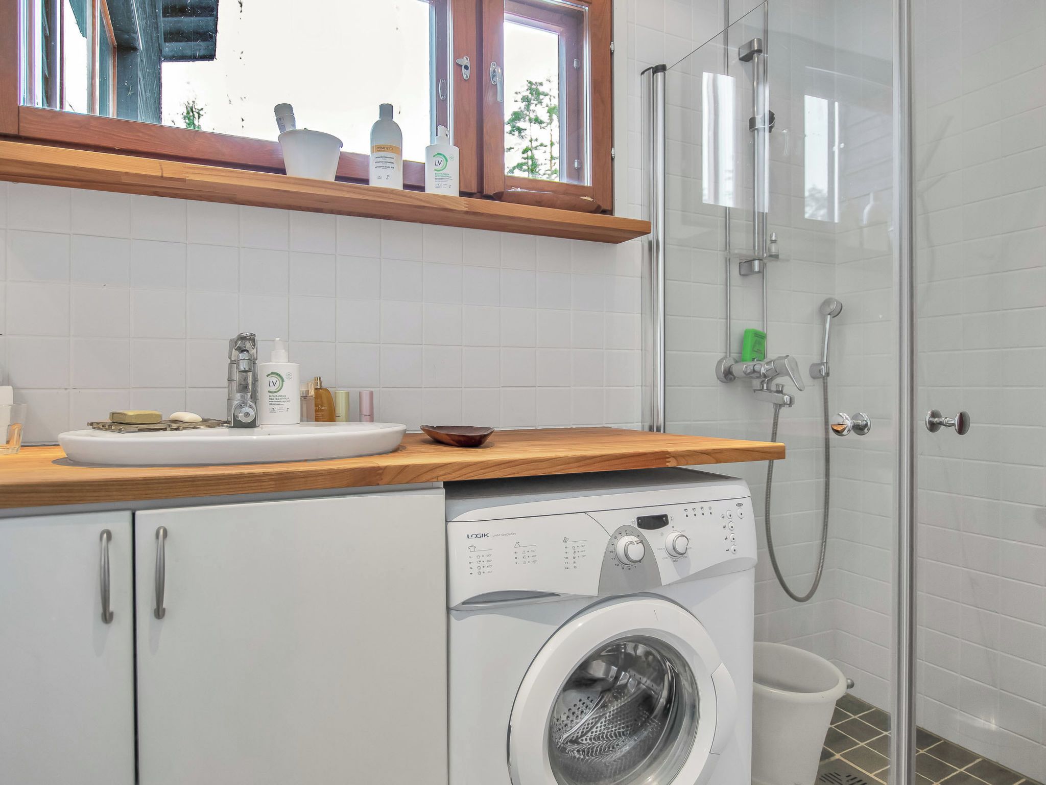 A modern bathroom with a washing machine, a glass shower enclosure, and a wooden countertop with toiletries.