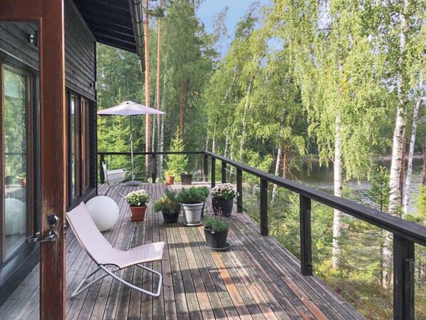 A wooden deck with a lounge chair, potted plants, and a view of a forest and water.