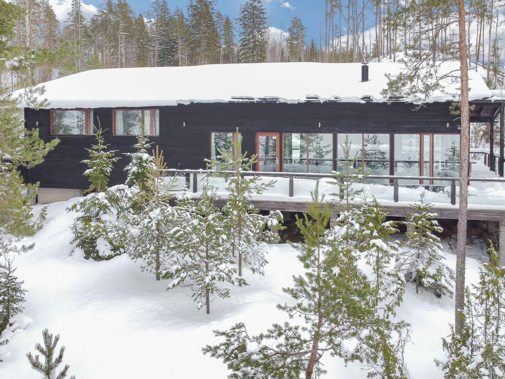 A modern cabin with a black exterior, large windows, and a deck surrounded by snow-covered trees.