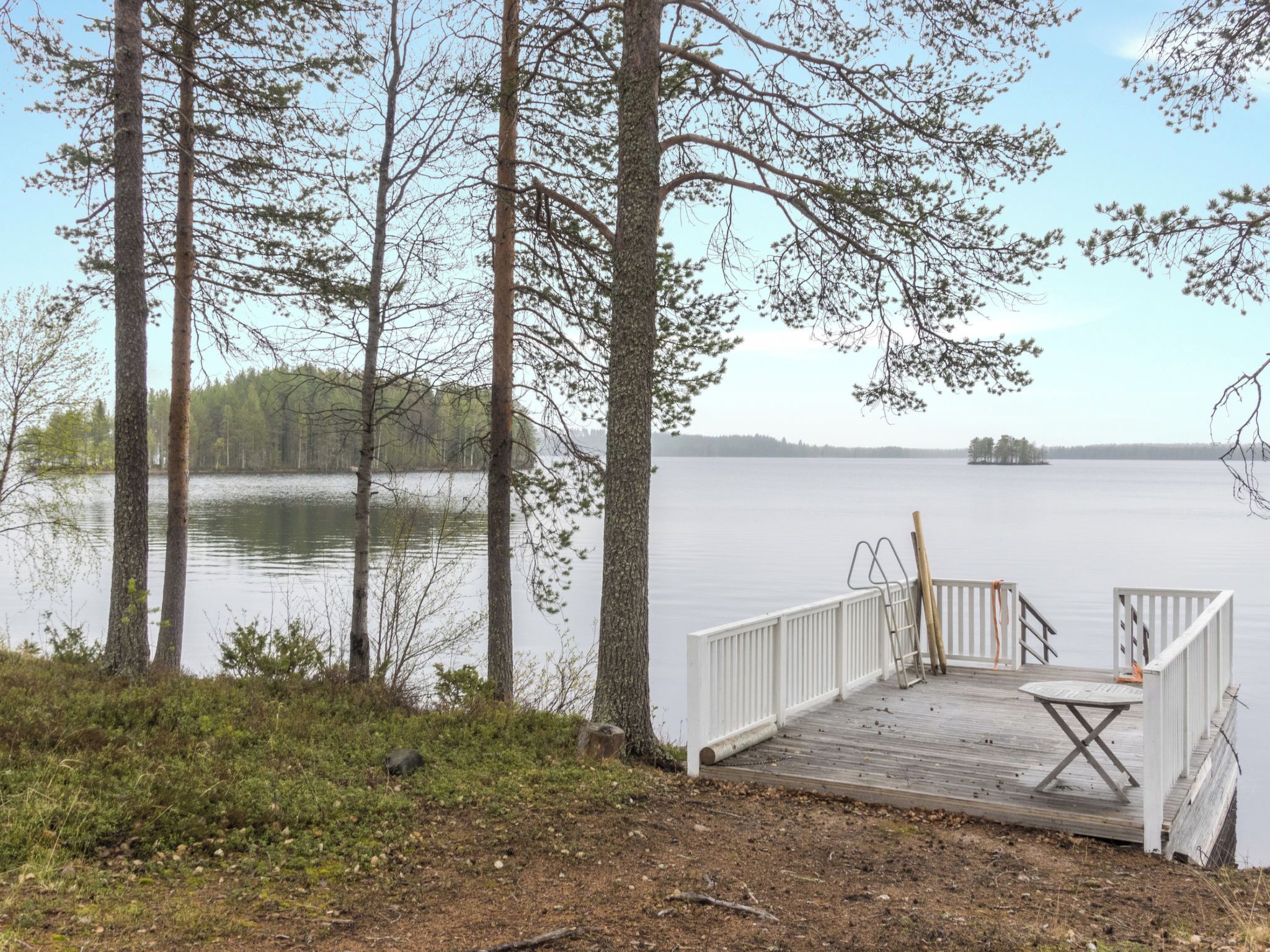 Eine Holzsteganlage mit weißer Geländer, einem Tisch und einem Stuhl, mit Blick auf einen ruhigen See, im Hintergrund Bäume.