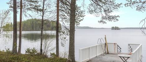 A wooden dock with a white railing, a table, and a chair, overlooking a calm lake with trees in the background.