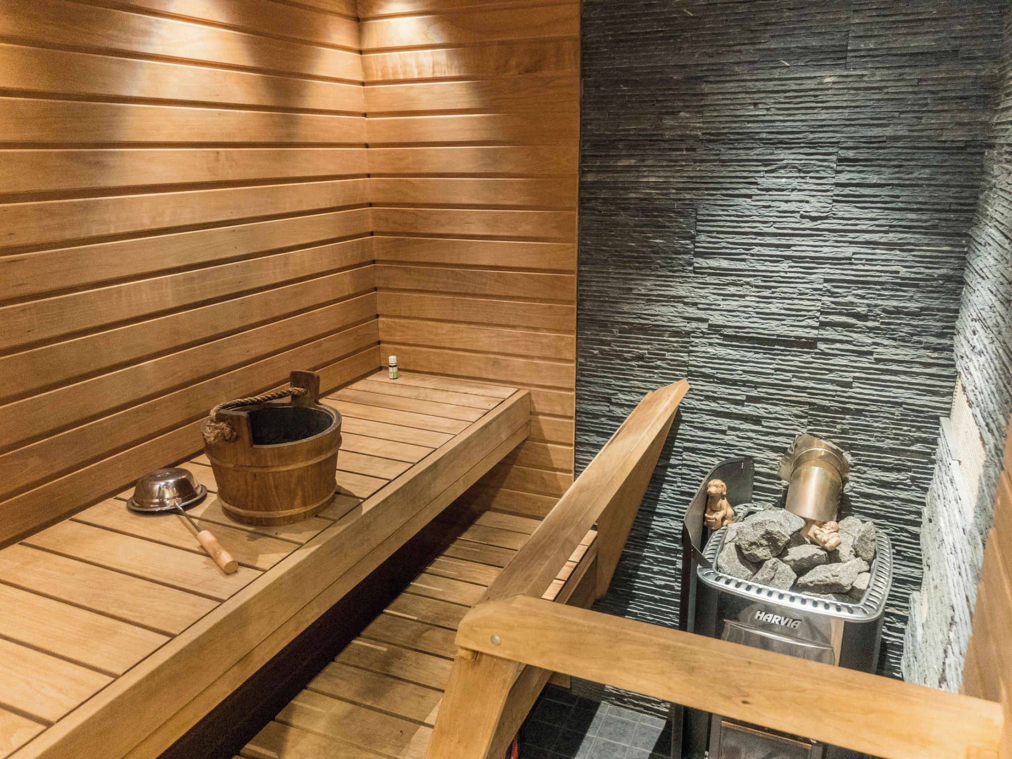 Una sauna con bancos de madera, un muro de piedra y un baño de vapor con un cabezal de sauna Harvia.