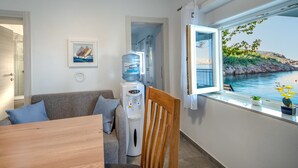 A room with a wooden table, a grey sofa with blue cushions, a water dispenser, and a window with a view of a rocky coastline. - Bajadera by Interhome (Jablanac)