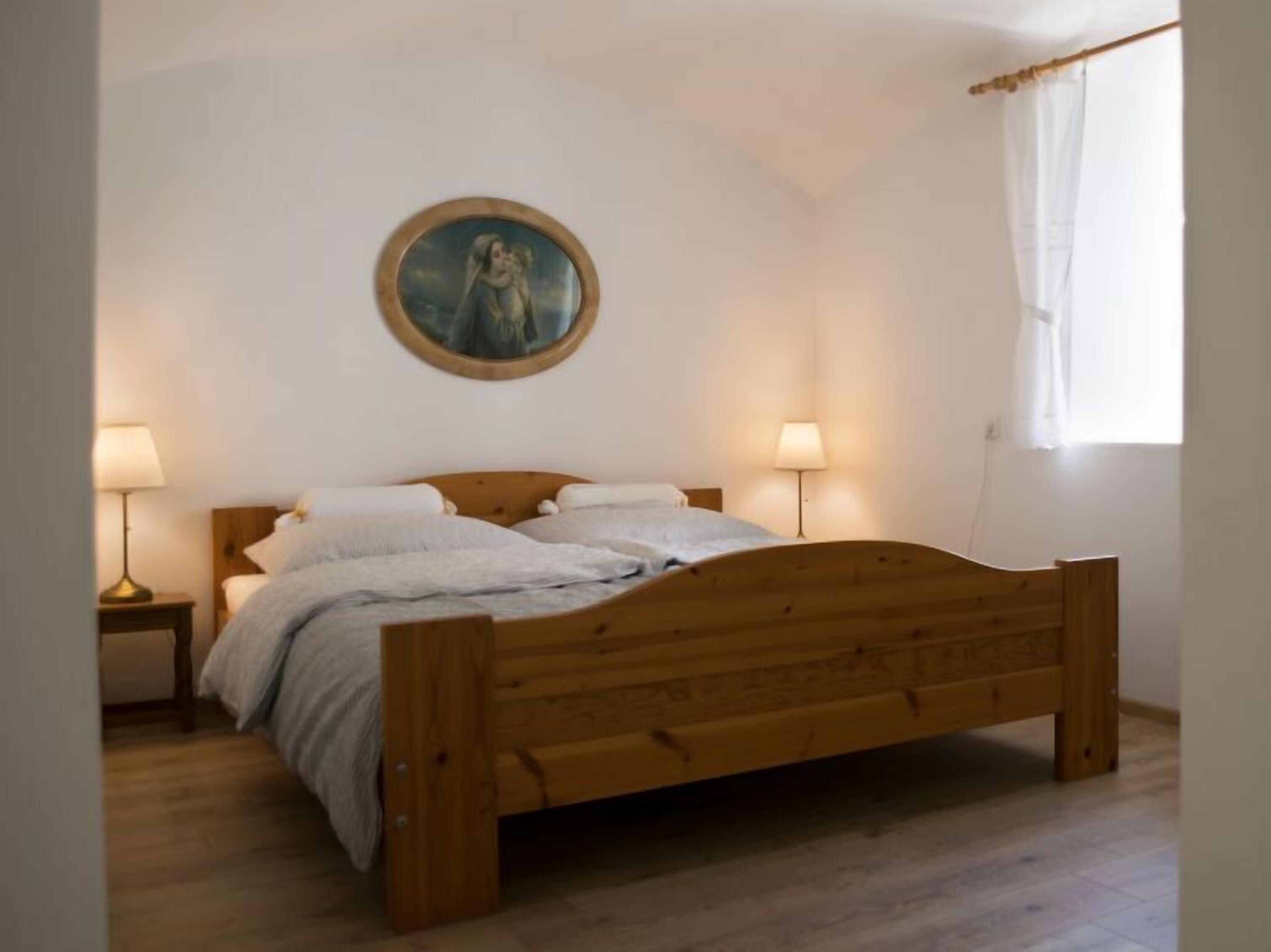 A bedroom with a wooden bed, two bedside lamps, a round painting on the wall, and a window with a white curtain.