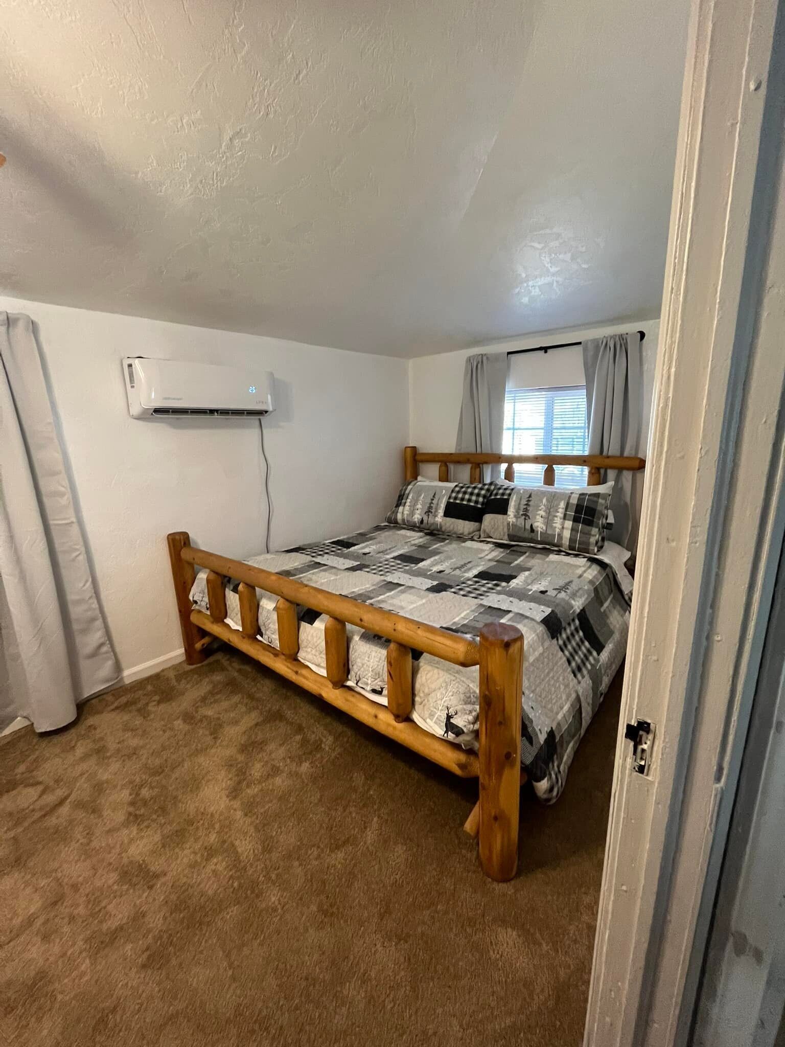 A bedroom with a wooden bed, a white wall air conditioner, and a window with curtains.