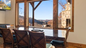 Un comedor con una mesa grande y sillas, vistas a montañas a través de amplios ventanales, y un edificio de piedra en el exterior.